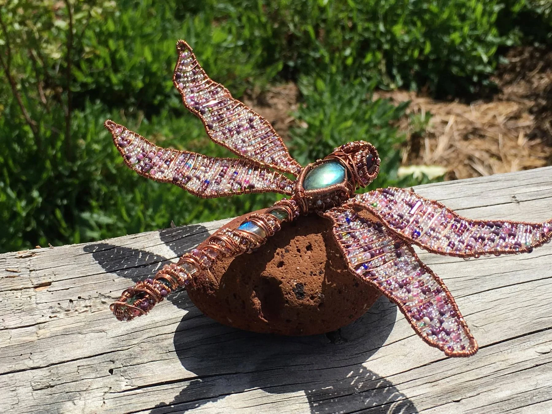 Handmade pink-beaded copper dragonfly resting on lava stone in sunlight.